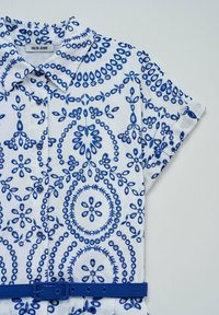 Camisa blanca con bordado floral y circular en azul, de mangas cortas, con botones en el frente y un cinturón azul a juego. Diseño casual.