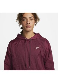Burgundska Nike jakna s kapuco z gubano teksturo, ki ima sprednji trak in majhen bel logo na levem prsnem delu.