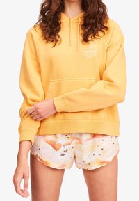 Sudadera de algodón amarilla con un bolsillo frontal y capucha con cordón. Combinada con unos pantalones cortos claros que presentan un patrón multicolor de montañas y pájaros.
