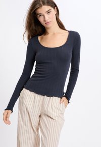 Młoda kobieta z długimi brązowymi włosami, ubrana w dopasowaną, granatową bluzkę z długim rękawem i luźne beżowe spodnie w paski, pozująca na białym tle.