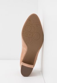 Semelle d’une chaussure habillée en daim beige avec des motifs en relief de pièce et de texte sur le dessous, reposant contre une surface d’angle blanche.
