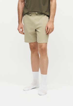 Férfi, bézs, zsinóros derékpántos rövidnadrágban, fehér bokazokniban és olivazöld pólóban, egyszínű fehér háttér előtt áll.