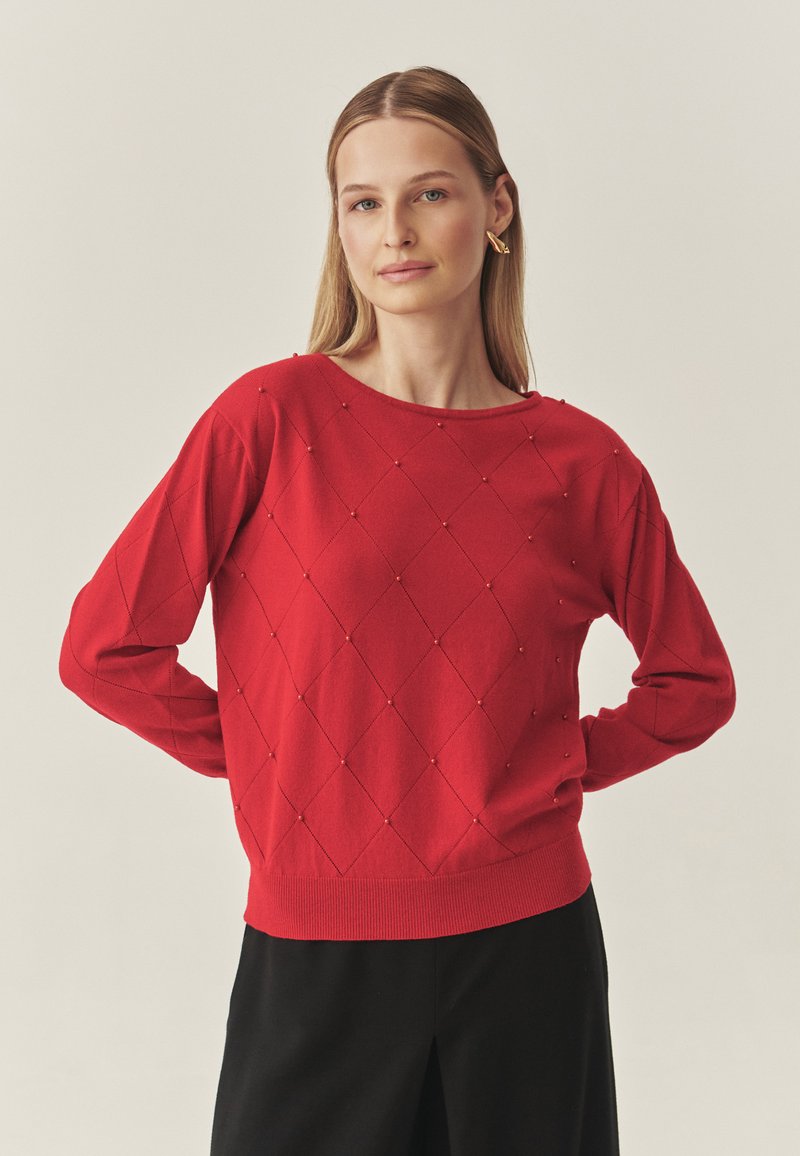 Piros kötött pulóver gyémántmintákkal és textúrált részletekkel. Kerek nyakkivágással és bordázott mandzsettákkal, valamint szegéllyel rendelkezik. Fekete nadrággal viselve.