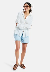 Witte longsleeve overhem met een subtiele textuur, lichtblauwe denimshorts en bruine slidesandalen. Bruine zonnebril en gelaagde kettingen.
