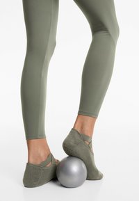Olajzöld leggings, sima textúrával, olajzöld hálós lábbelivel, keresztbe fűzött pántokkal, egy ezüst edzőlabdán pihen.
