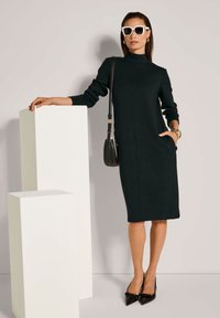 Schwarzes langärmliges Kleid mit hohem Kragen, Seitentaschen und geradem Schnitt. Getragen mit eleganten schwarzen Stöckelschuhen und übergroßen weißen Sonnenbrillen.