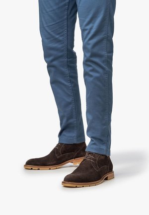 Botas de ante marrón hasta el tobillo con detalles de cordones, combinadas con pantalones ajustados azul claro, que muestran una textura suave y un color de suela contrastante.