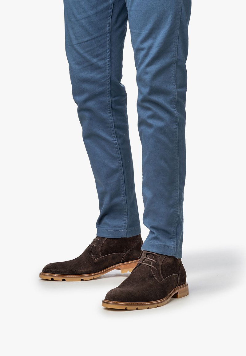 Stivali alla caviglia in suede marrone con dettagli in pizzo, abbinati a pantaloni aderenti blu chiaro, che mostrano una superficie liscia e un colore della suola in contrasto.