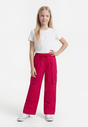 Young girl standing with one hand on hip, wearing a white short-sleeve shirt, bright pink cargo pants, and white sneakers.