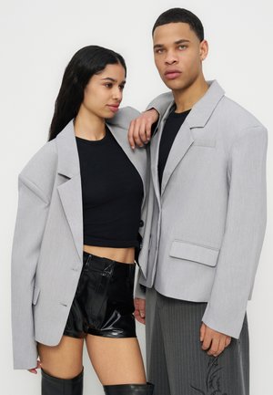 Two models wearing oversized light gray blazers; one pairs it with black crop top, vinyl shorts, and thigh-high boots, the other with pinstripe pants.