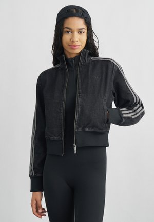 Jeune femme portant une veste noire courte zippée avec des rayures argentées sur les manches et une casquette noire portée à l'envers, debout devant un fond uni.
