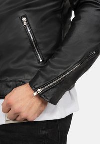 Zwarte leren jas met zilveren ritsen, voorzien van een getailleerd ontwerp, een soepele textuur en ritsmanchetten. Een hand houdt een kaart dicht bij de tailleband.