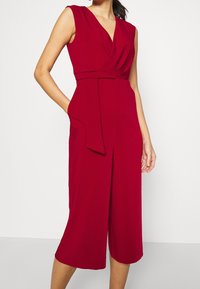 Combinaison sans manches rouge avec un profond décolleté en V, ceinture à la taille, jambes larges et poches latérales ; fabriquée en tissu lisse avec un drapé subtil.