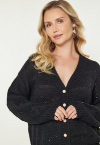 Cardigan nero a maglia con scollo a V, decorato con bottoni perlacei e tessuto testurizzato. Maniche lunghe, leggermente a sbuffo, con polsini aperti.