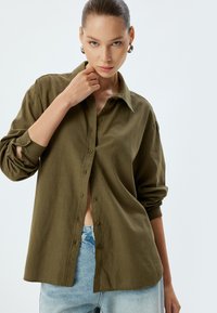 Camicia verde oliva con bottoni in tessuto morbido, caratterizzata da un colletto, maniche arrotolate e una vestibilità rilassata, abbinata a jeans azzurri chiari.