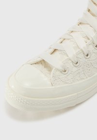 Fehér csipkés sneaker fényes gumitalppal, selyemfűzőkkel és textúrált csipke részletekkel a felsőrészen. Gumitalp szőtt mintázattal.