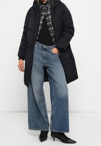 Sort puffcoat med hætte, lagdelt over en sort top og ternet skjorte. Lys til mørkeblå jeans med vide ben og sorte støvler med hæl.