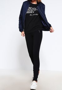 Persona que lleva una chaqueta azul marino, leggings negros, zapatillas blancas y una camiseta negra con el texto "REALMENTE NECESITO ZAPATOS NUEVOS - Yo, todos los días."