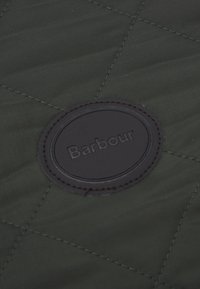 Πράσινο καπιτονέ ύφασμα με έναν στρογγυλό μαύρο δερμάτινο θησαυρό που έχει το "Barbour" ανάγλυφο στο κέντρο, περιτριγυρισμένο από μια ραμμένη στεφάνη.