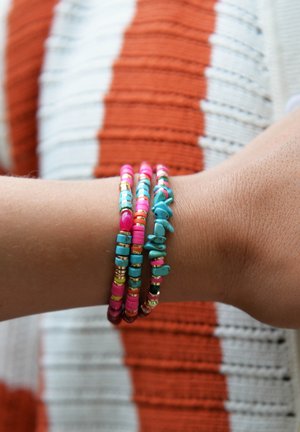 Bracelets colorés en perles roses, turquoise et dorées, présentant des formes et des tailles variées, portés sur un poignet contre un fond texturé.