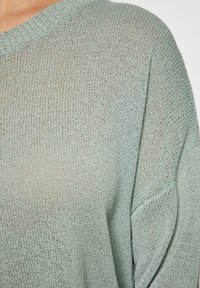 Pulover tricotat verde deschis, cu o croială lejera, guler lat cu nervuri și un model texturat. Materialul pare moale și respirabil.