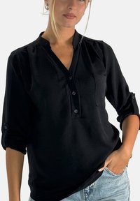 Zwarte blouse met knopen, een V-hals, lange mouwen opgerold tot de elleboog, een borstzak en een zachte, gladde textuur.