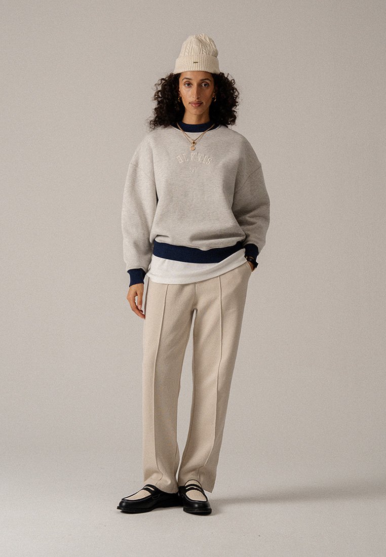 Szürke, túlméretezett pulóver tengerészkék díszítésekkel, krémszínű, bő szárú nadrág és fekete cipő fehér részletekkel. A modell texturált kötött sapkát visel.