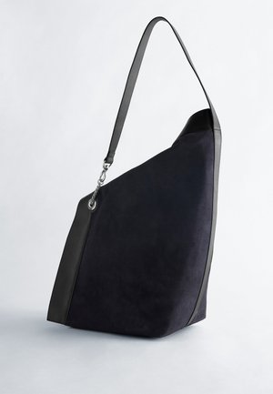 Grand sac à épaule en daim noir avec une seule bandoulière en cuir ajustable et une quincaillerie métallique argentée sur un fond uni.