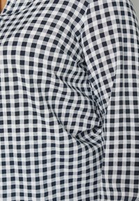Kangas berwarna hitam dan putih yang dicetak kotak-kotak membentang di atas tubuh, menunjukkan lipatan dan kontur pas badan pada bagian atas torso.