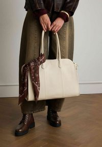 Crème lederen handtas met gestructureerde vorm, dubbele handvatten en een bruine sjaal met patroon vastgebonden aan het handvat. Model draagt bruine veterschoenen en groene broeken.