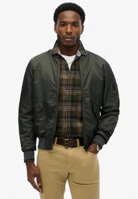 Donkergroen bomberjack met ritssluiting aan de voorkant, met ribgebreide manchetten. Draagt over een flanellen shirt met een groen en oranje ruitpatroon en een lichtblauwe kraag.