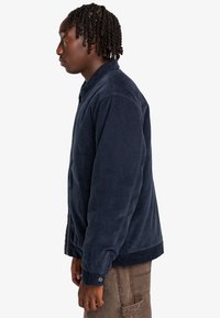 Navy corduroy jacket with a buttoned front, side pockets, and a relaxed fit. Features a collar and ribbed cuffs.