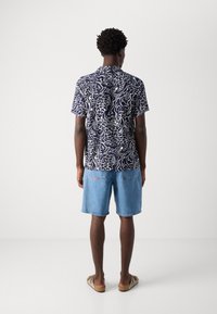 Camisa de manga curta azul-marinho com padrão abstrato branco, combinada com calções de ganga de lavagens claras. Usada com sandálias castanhas. Design simples, ajuste casual.