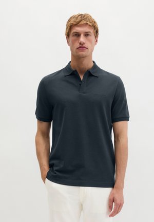 Homme portant un polo à manches courtes gris foncé et un pantalon blanc, debout devant un fond clair uni.