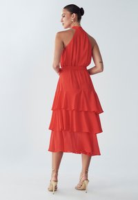 Red tiered dress made of lightweight fabric, featuring a halter neck, belted waist, and ruffled layers, paired with gold strappy sandals.