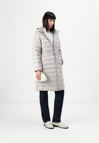 Lys grå dunjakke med hette, belte i midjen og horisontalt quiltet. Kombinert med mørke jeans og lyse slip-on-sko, mens hun holder en liten veske.