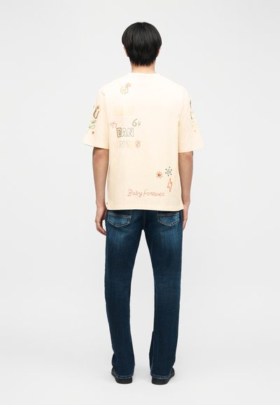 Άτομο με κοντά μαύρα μαλλιά στέκεται γυρισμένο προς τα πίσω, φορώντας ανοιχτό μπεζ πουκάμισο με πολύχρωμο κεντητό κείμενο και σκούρο μπλε τζιν.