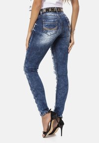 Dunne blauwe denim jeans met een gescheurde afwerking aan de zijkanten, voorzien van achterzakken en een zwarte gesp met studs. Zwarte hakken.