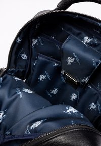 Svart ryggsäcksinteriör med en slät tygfoder som har ett marinblått mönster av små, vita U.S. Polo Assn.-logotyper och fack.
