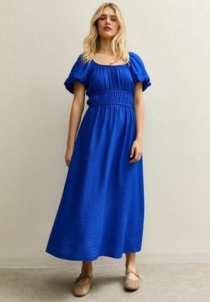 Vestido maxi azul con mangas abullonadas, cintura fruncida y tela texturizada. Llevado con zapatos planos beige, sobre un fondo neutro.