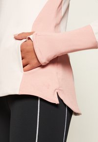 Lehký sportovní top s růžovými a bílými barevnými bloky, vysokým límcem, bočními rozparky a logem na spodním lemu. Šikmé rukávy s otvory na palce.