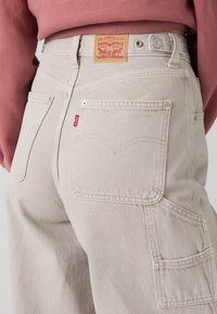 Πίσω όψη ανοιχτό μπεζ τζιν Levi's με δερμάτινη ετικέτα μάρκας, κόκκινη ετικέτα και πολλαπλές τσέπες, φορεμένα με ροζ μπλούζα μακρυμάνικη.
