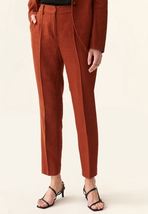 Pantaloni in lino color ruggine con una vestibilità sartoriale, caratterizzati da pieghe frontali e una texture liscia. Abbinate a tacchi neri con fasce.