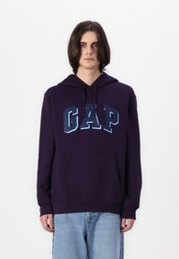 Felpa viola con tasca a marsupio, caratterizzata da un grande logo "GAP" in tonalità sfumate di blu sul davanti. Tessuto morbido e vestibilità comoda.