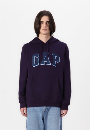 Ljubičasti dukserica s kenguruvim džepom, s velikim "GAP" logotipom u gradijentnim nijansama plave boje sprijeda. Mekana tkanina i opušteni kroj.