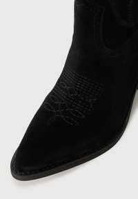 Bottine en suède noir avec un bout pointu, des coutures détaillées dans un design à motifs, et une semelle en cuir lisse.