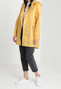 Gul regnjacka med huva, prydd med små, flerfärgade regndroppe-mönster. Har elastiska ärmslut och dragsko i midjan.