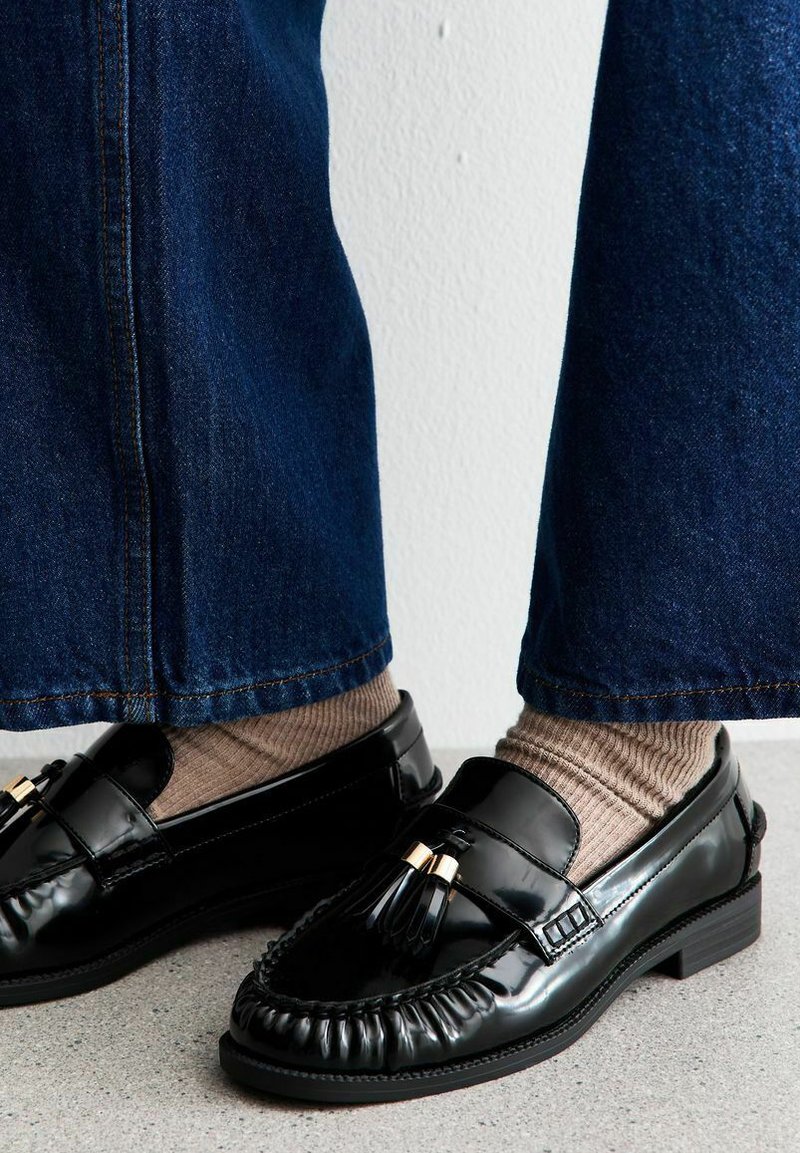 Mocassini in vernice nera con nappine, caratterizzati da un design cucito, abbinati a calzini beige a coste e pantaloni di denim a gamba larga.