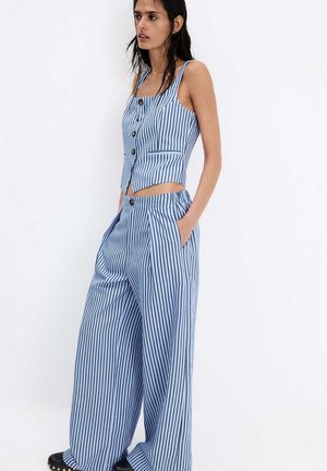 Mujer con top sin mangas de rayas verticales azules y blancas y pantalones de pierna ancha con zapatos negros, de pie sobre un fondo blanco liso.