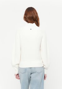 Kvinne med langt rødt hår iført en hvit strikkegenser og lyseblå jeans, vist fra baksiden mot en hvit bakgrunn.